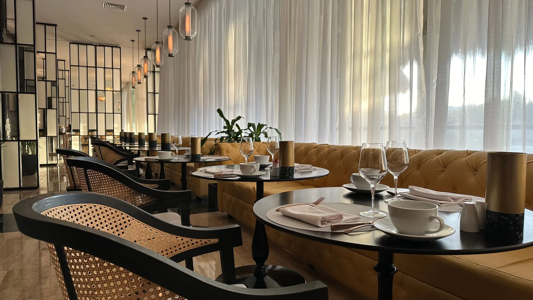A restaurant dining room set for service, with cane chairs, tufted banquettes and pendant lights